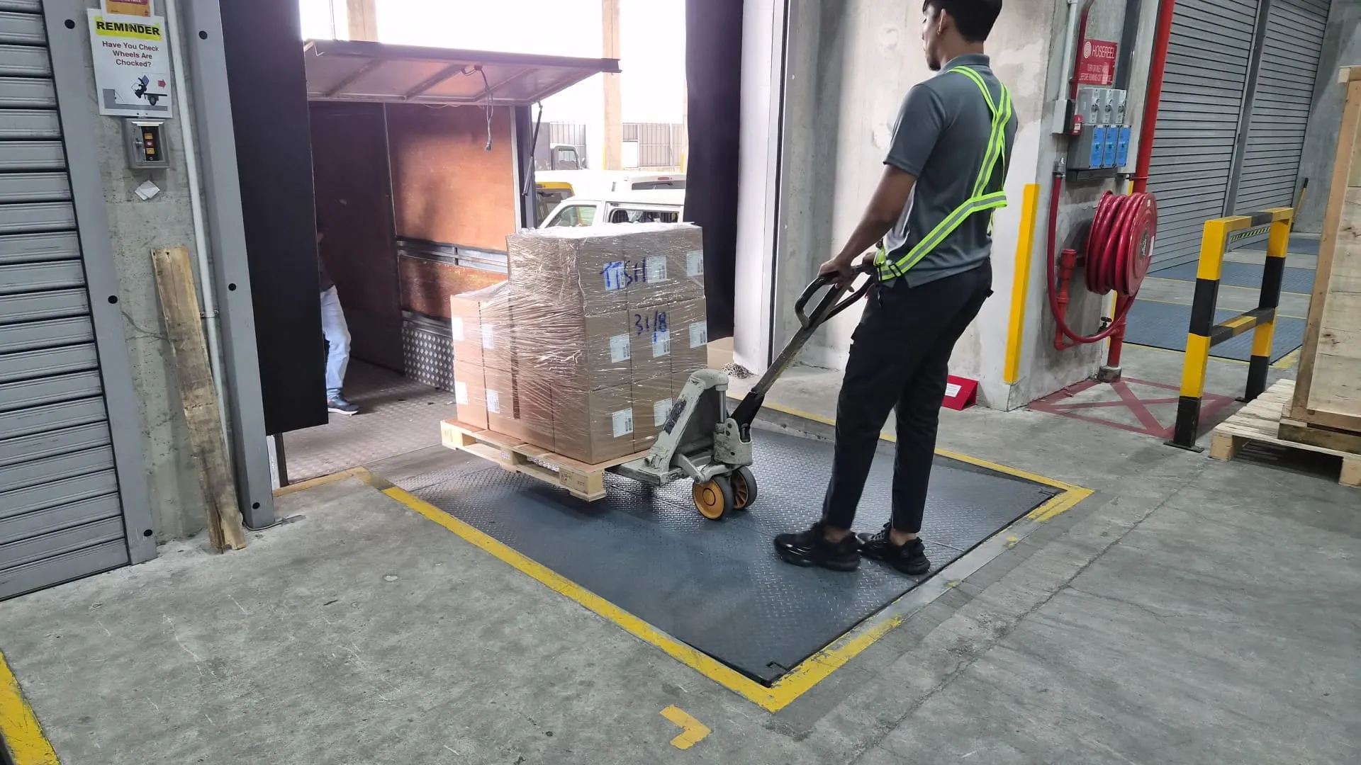 Driver seen from behind moving a pallet of cartons on a pallet jack across a dock plate towards an open lorry tailgate, red fire-hose reel on the wall
