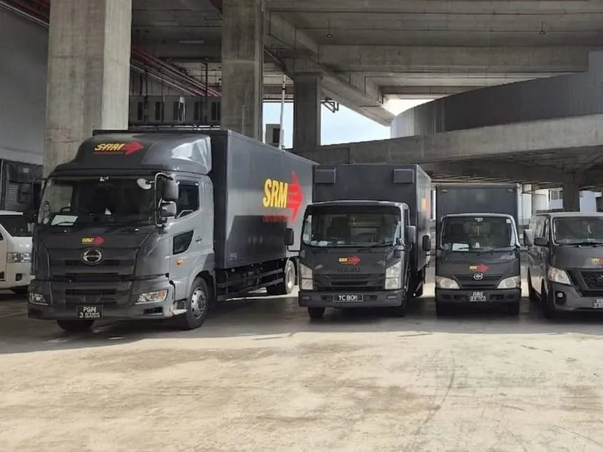 SRM Logistics 24-ft Hino box lorry and an Isuzu box lorry parked in the company yard beneath a concrete flyover, the yellow SRM wordmark and red arrow printed along the side of the Hino's box body