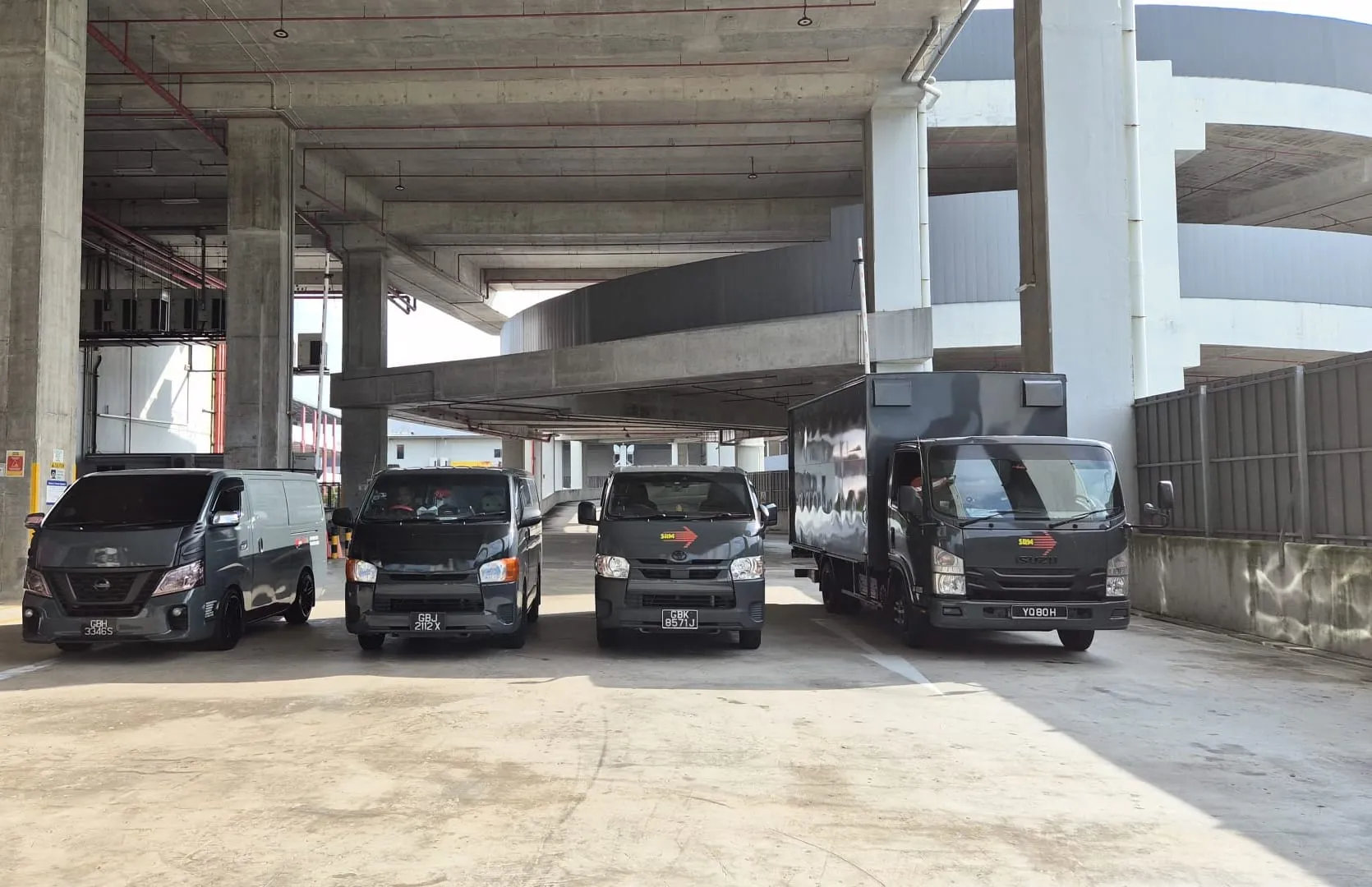 Four SRM Logistics vehicles in a row: a Nissan NV350 van, two Toyota Hiace vans and an Isuzu box lorry, all in dark grey livery
