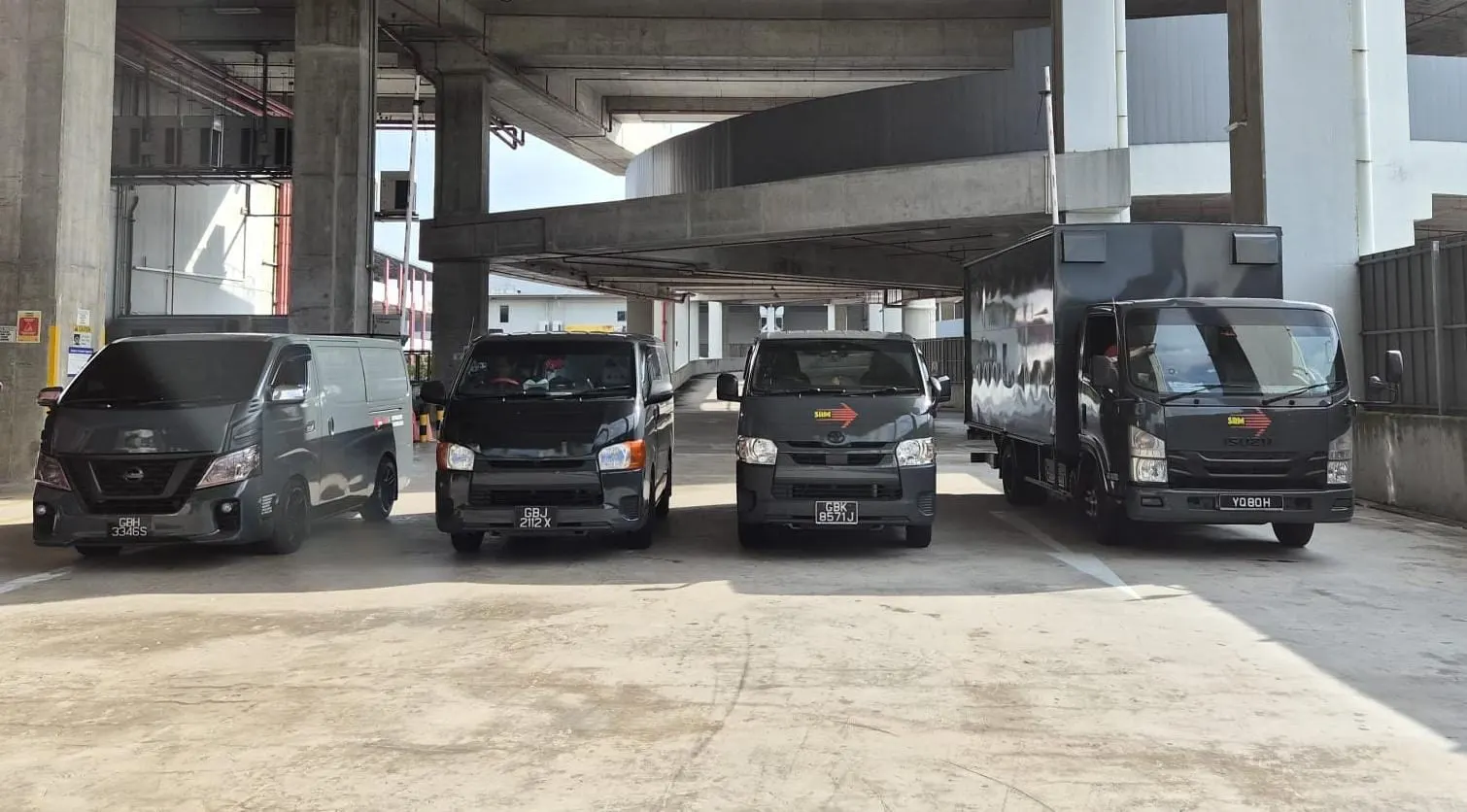 Close front view of four dark grey SRM Logistics vans and a box lorry parked under a concrete flyover