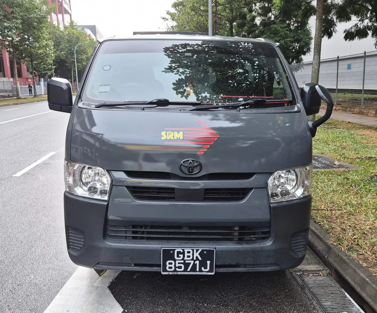 Front view of a dark grey SRM Logistics Toyota Hiace van parked at a Singapore roadside, the yellow SRM wordmark and red arrow on its bonnet